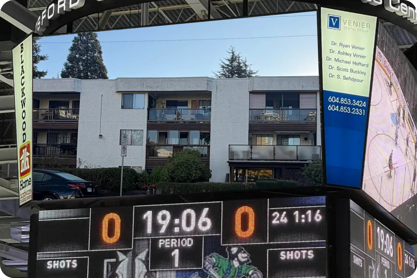 An arena jumbotron showing a three storey White Rock condo building with balconies, above a hockey scoreboard reading nil to nil in the first period