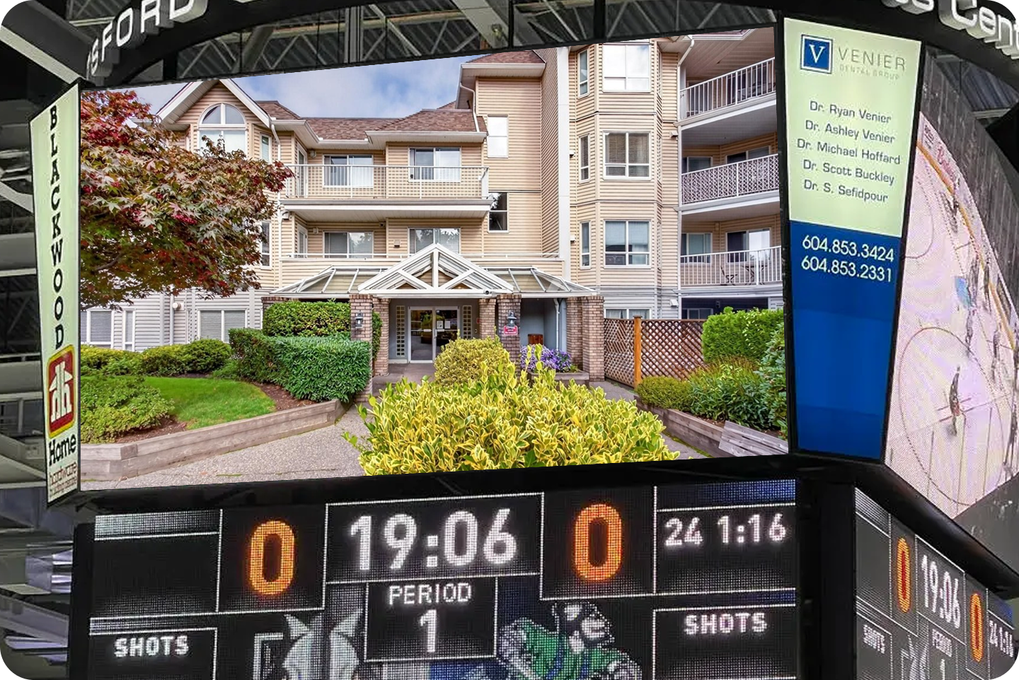 An arena jumbotron showing a beige four storey Whalley condo building with balconies and a landscaped entrance, above a hockey scoreboard reading nil to nil in the first period